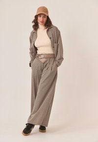 Mujer vestida con un suéter de cuello alto beige, chaqueta de cuadros y pantalones de pierna ancha, gorra marrón, cinturón ancho y zapatillas negras frente a un fondo liso.