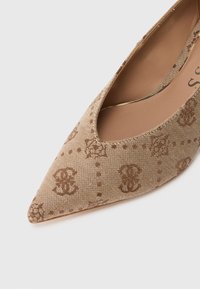 Spetsig beige tygsko med bruna broderade geometriska och blommotiv samt en slät tan färgad innersula mot en vit bakgrund.