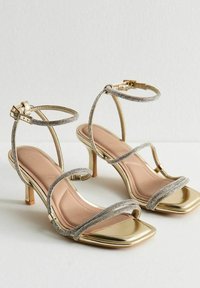 Goldene High-Heelsandalen mit quadratischen Zehen, dünnen Strassriemen, die über den Fuß verlaufen und sich mit einer Schnallenverschluss um den Knöchel wickeln.