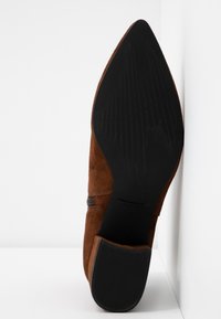 Botte en daim marron à bout pointu avec semelle noire texturée et talon bloc, photographiée par dessous sur fond blanc.