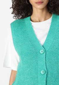 Turquoise knitted vest with a v-neck, featuring textured ribbing, two light blue buttons, worn over a white t-shirt.