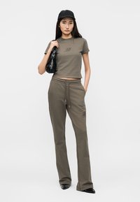 Νεαρή γυναίκα που φοράει πράσινο ελαιί crop top και αντίστοιχο παντελόνι φόρμας, μαύρο καπέλο, τσάντα ώμου και μαύρα μυτερά παπούτσια, όρθια.