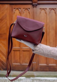 Burgundská crossbody taška vyrobená z texturované kůže, s klopovým uzávěrem, stříbrným kováním a nastavitelným popruhem. Ozdobena akcentem s logem.