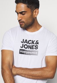 White short-sleeve t-shirt featuring bold black printed text: "JACK & JONES" and "PERFORMANCE." Soft fabric with a relaxed fit.
