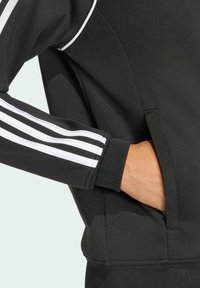 Giacca sportiva nera con strisce bianche sulle maniche, polsini a coste e una tasca laterale, realizzata in un tessuto liscio e strutturato.