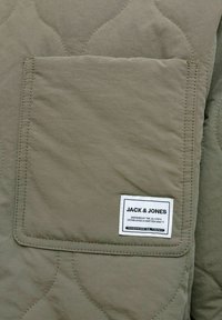 Gequilt stof van een olijfgroene jas met een vierkante borstzak met witte labeltekst, zichtbare stiksels en subtiele textuurpatronen.