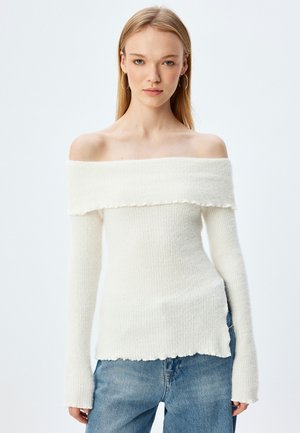 Off-shoulder witte geribde trui met rafelige randen, lange mouwen en een aansluitend ontwerp, gecombineerd met lichtblauwe spijkerbroek.
