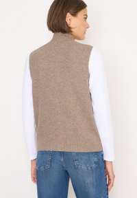 Beige Strick-Sweatervest mit hohem Kragen und geripptem Saum, getragen über einem weißen Langarmshirt, kombiniert mit blauen Jeans.