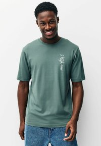 Halványzöld, rövid ujjú póló puha anyagból, fehér grafikával és szöveggel a bal mellrészen. Kényelmes szabás, kerek nyakkivágással.