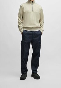 Ljusbeige zip-up tröja med hög krage, kombinerad med mörkblå denim cargobyxor med sidofickor och avslappnad passform. Svarta skor fulländar looken.