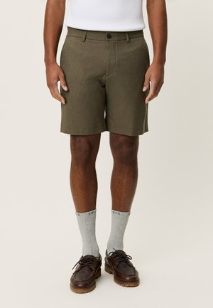 Krótkie spodenki oliwkowe wykonane z teksturowanej tkaniny, wyposażone w przednie zapięcie na guziki i boczne kieszenie, połączone z szarymi skarpetami i brązowymi butami.