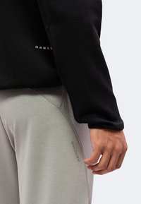 Άτομο με ανοιχτό γκρι παντελόνια Oakley και μαύρο μακρυμάνικο μπλουζάκι Oakley, χέρι χαλαρό στο πλάι.