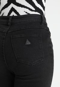 Svarta denimbyxor med en standard bakficka och ett triangulärt logopatch samt en hög midja. Texturen ser slät ut.