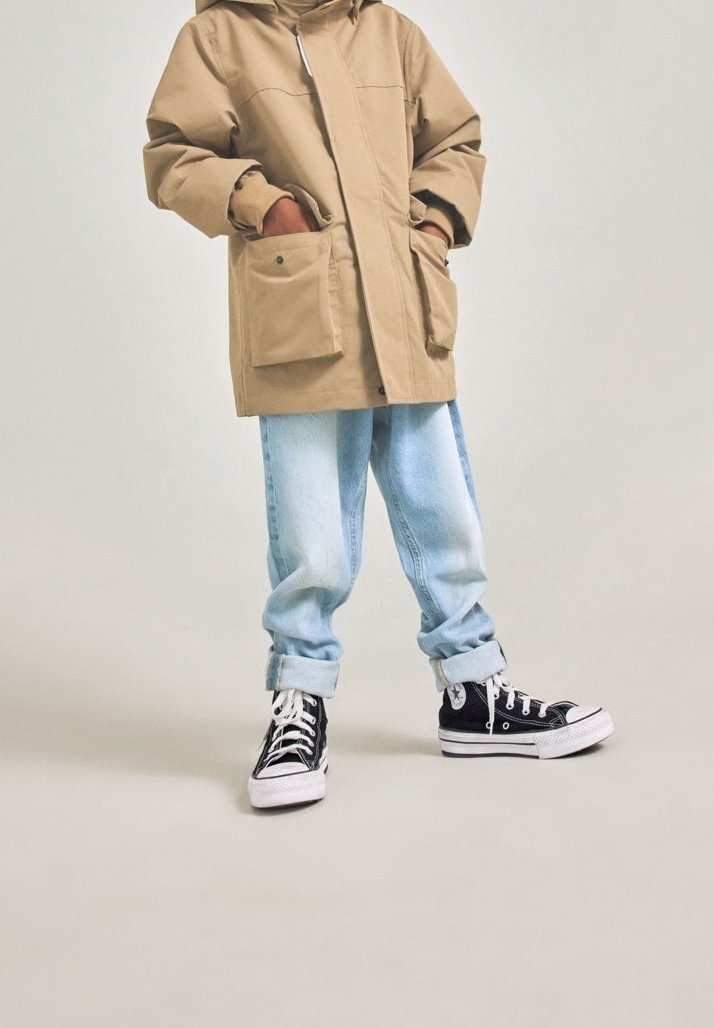 Personne portant une parka beige avec de grandes poches, un jean bleu clair retroussé et des baskets montantes noires sur un fond clair uni.