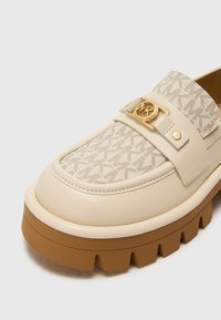 Beige mokasīni ar masīvu brūnu zoli, rakstainu auduma paneļa un zelta apļveida MK logo sprādzi uz ādas joslas pāri virsotnei.