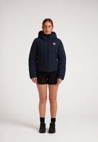 Giacca puffer blu navy con cappuccio, cerniera frontale e patch con logo. Presenta una texture trapuntata, maniche lunghe e tasche laterali.