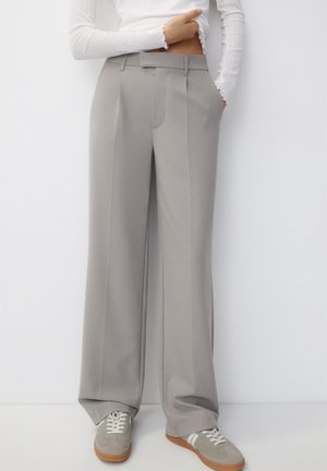 Personne portant un pantalon gris taille haute à jambes larges avec des plis sur le devant, associé à des baskets grises et blanches, debout contre un mur blanc.