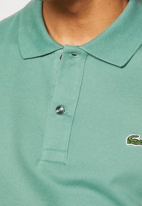 Polo vert clair en tissu texturé. Comprend un col replié, une patte de boutonnage à deux boutons et un petit logo brodé dans un coin.