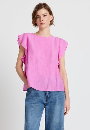 Frau trägt eine locker sitzende, ärmellose rosa Bluse mit Rüschen an den Schultern und blaue Jeans, steht mit einer Hand in der Tasche.