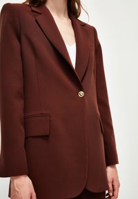 Bruine op maat gemaakte blazer met een notch revers, sluiting met één gouden knoop en twee voorzakken. Gladde textuur, minimale details.