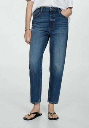 Personne portant un jean bleu droit et des sandales noires avec des accents dorés, debout avec une main dans la poche sur fond uni.