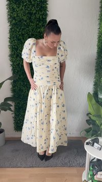 Abito giallo floreale con maniche a sbuffo, corpetto arricciato e gonna a strati. Presenta motivi floreali blu e un design fluido e tessurizzato.