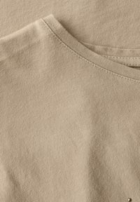 Tejido de algodón beige con una textura suave y un escote redondo. Presenta un sutil detalle de costura y un acento decorativo cerca del dobladillo.
