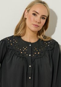 Svart blus med en scallopad ögonhål broderad krage och knappstängning fram. Mjukt tyg med subtil struktur och veckade detaljer.