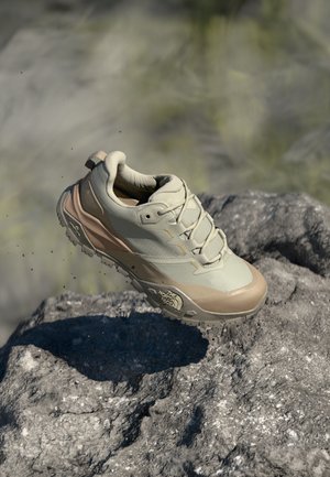 Beige idrettssko med en teksturert overdel, forsterket tåhette og robust såle. Snøringer og reflekterende detaljer er synlige mot en steinete bakgrunn.