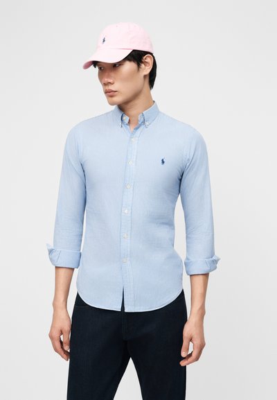 Férfi világoskék csíkos ingben, felhajtott ujjakkal és rózsaszín baseballsapkában, egyszínű háttér előtt áll.