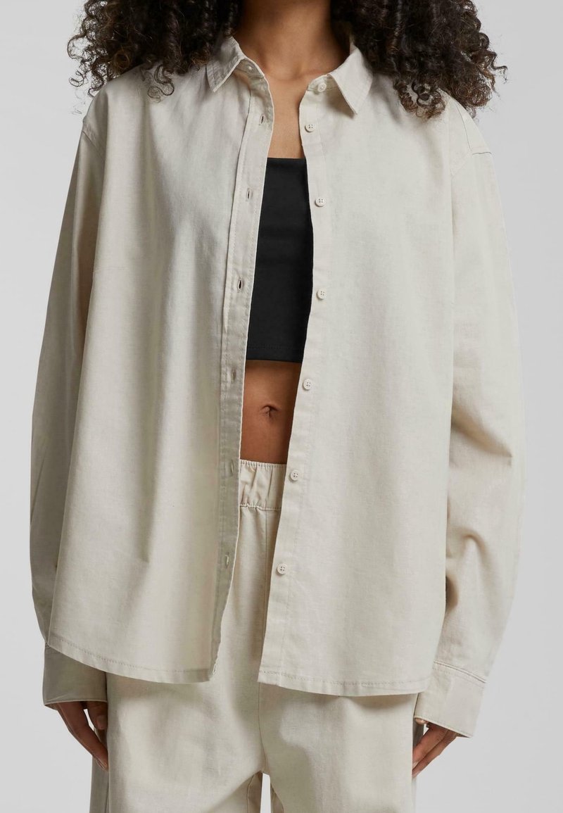 Femme portant une chemise beige oversize boutonnée sur un crop top noir et un pantalon beige assorti avec taille élastique.