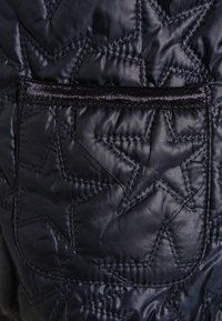 Tessuto nero trapuntato con motivi a stella, caratterizzato da una piccola tasca liscia e una texture lucida; include un bordo nero attorno alla tasca.