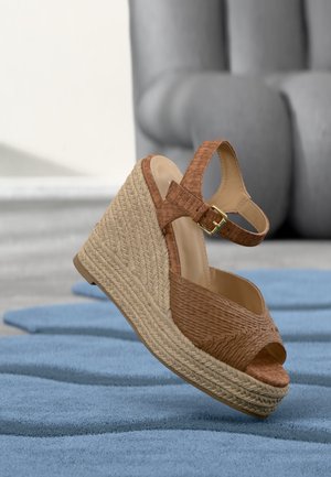 Sandale compensée marron en tissé avec sangle à la cheville et plateforme en jute tressé sur tapis bleu, canapé gris flou en arrière-plan.