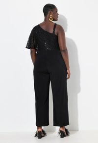 Mono negro con cuerpo de encaje y lentejuelas, diseño asimétrico y silueta de pierna ancha. Tacones negros ajustados con detalles de plumas.