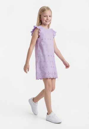 Noor tüdruk kõnnib, kandes varrukateta lavendlivärvi kleiti silmkoeliste detailidega ja valgeid tenniseid, naeratades, valgel taustal.