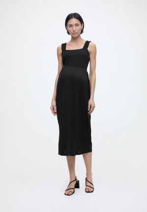 Donna incinta con capelli corti e scuri che indossa un vestito midi senza maniche nero e tacchi neri con cinturini, in piedi davanti a uno sfondo bianco.