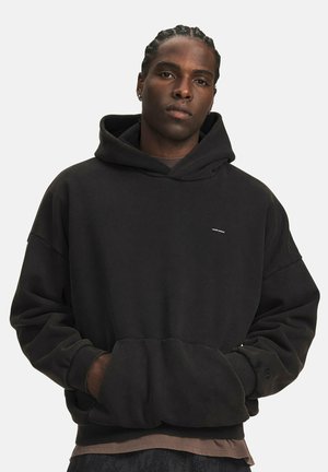 Jonge man met gevlochten haar, draagt een losse zwarte hoodie met handen in de voorzak, staand tegen een effen lichte achtergrond.