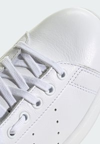 Bílé kožené tenisky s zaoblenou špičkou, texturovaným povrchem a bílými tkaničkami. Obsahují perforované detaily a kulaté očka po stranách.