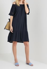 Marinblå off-the-shoulder klänning med volangärmar, samlad midja och lager på lager nederdel. Slätt tyg. Kombinerad med ljusblå sandaler med öppen tå.