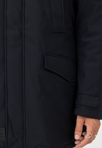 Schwarzer Parka mit glatter Textur, ausgestattet mit einer Brusttasche und Handwärmer-Taschen, und einem silbernen Ring, der an der Hand sichtbar ist.