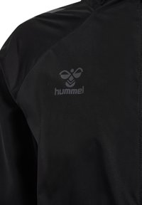 Detail einer schwarzen Sportjacke mit dem hummel-Logo auf der Brust, einem Bienen-Symbol und Chevron-Muster.