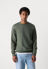 Olivenfarvet sweatshirt med rund halsudskæring og lange ærmer. Har et lille logo-patch på brystet og ribbede manchetter. Båret over lyse jeans.