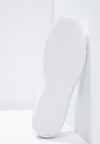 Semelle d'une basket blanche avec un motif à chevrons posée contre un coin blanc entre un mur et le sol.