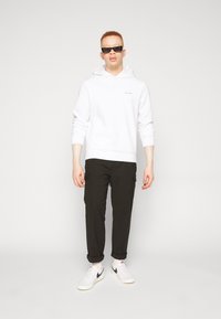 Sweatshirt à capuche blanche avec logo Calvin Klein, pantalon cargo noir et sneakers blanches avec accents noirs ; le mannequin porte des lunettes de soleil noires.