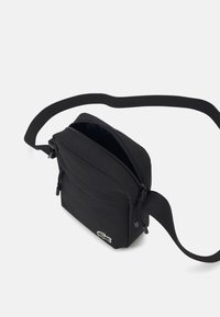 Černá crossbody taška vyrobená z odolné tkaniny, s zipovým uzavíráním, nastavitelným popruhem a malou zelenou nášivkou. Kompaktní obdélníkový tvar.
