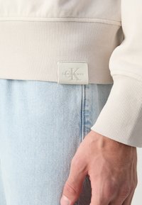 Ourlet beige côtelé de sweat-shirt avec patch logo Calvin Klein Jeans au-dessus d'un jean denim bleu clair et une main détendue sur le côté.