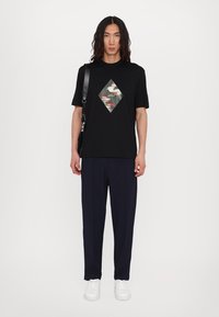 Tricou negru cu o patch în formă de diamant în camuflaj, asortat cu pantaloni navy și adidași albi, purtat de un model cu păr lung.