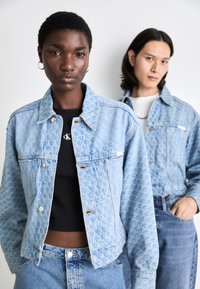 Chaqueta de denim azul claro con textura estampada, con cuello, botones frontales y bolsillos laterales. Usada sobre una blusa negra.