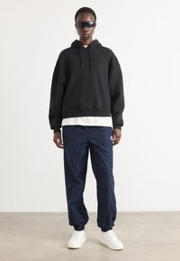 Zwarte hoodie, witte gelaagde shirt, marineblauwe trainingsbroek en witte sneakers. De outfit heeft een ontspannen fit en minimale versieringen. Zwarte zonnebril gedragen.