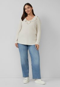 Hellbeige, geripptes Sweatshirt mit V-Ausschnitt und Volantbesatz, kombiniert mit weit geschnittenen blauen Jeans und weißen Turnschuhen. Glatte Textur, lässige Passform.
